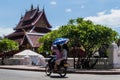 Two Laotian girls ride a scooter in front of the Wat Mai Suwannaphumaham temple in Luang Prabang, Laos, holding an umbrella
