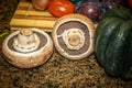 Two large button mushrooms sit on a marble cabinet with cuttying board and acorn squash and other veggies