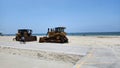 Two large rusty yellow bulldozers parked in the sand at Rosie\'s Dog Beach with blue ocean water and people in the sand