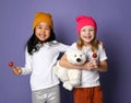 Two laughing kid girls in white t-shirt and colorful hats hold polar bear toy and lollipop candy on purple Royalty Free Stock Photo