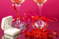 Two legs of champagne glasses tied with red ribbons on a pink background next to a ring in a box a candle in a heart candlestick