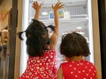 Little Asian baby girls, sisters, looking and touching the bakery fridge as they were irresistibly hungry for beautiful cakes