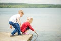 Two little blonde kids, boy and girl, sitting on a pier on a lake or river and searching for something or fishing with wooden Royalty Free Stock Photo