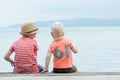 Two little boys sit on a pier, against the sea and mountains. Ba