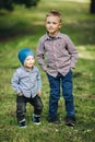 Two little boys wearing in casual looks posing in park holding hands in pockets.