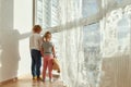 Two little children in home clothes holding hands while standing indoors and looking out of a large window on a sunny Royalty Free Stock Photo