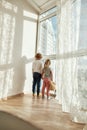 Two little children in home clothes holding hands while standing indoors and looking out of a large window on a sunny Royalty Free Stock Photo