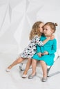 Two little girlfriends in the identical dresses of different colors sitting on a chair in a studio with white walls
