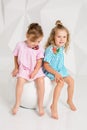 Two little girlfriends in the identical dresses of different colors sitting on a chair in a studio with white walls