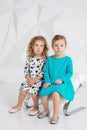 Two little girlfriends in the identical dresses of different colors sitting on a chair in a studio with white walls