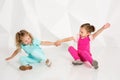 Two little girlfriends in the identical overalls of different colors sitting on the floor in a studio with white walls