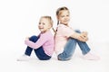 Two little girlfriends in jeans and pink sweaters are sitting and smiling. White background