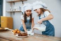 Two little girls in blue chef uniform pouring milk into glasses on the kitchen with cookies on table Royalty Free Stock Photo