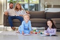 Two little girls doing homework lying on living room floor with their parents relaxing on couch. Little kids using Royalty Free Stock Photo