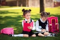 Two Little school girls with pink backpack sitting on grass after lessons and thinking ideas, read book and study Royalty Free Stock Photo