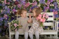 Little twin girls in the garden on a bench against the background of a floral wall with bouquets of flowers in their hands Royalty Free Stock Photo