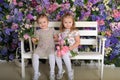 Little twin girls in the garden on a bench against the background of a floral wall with bouquets of flowers in their hands Royalty Free Stock Photo