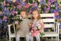 Little twin girls in the garden on a bench against the background of a floral wall with bouquets of flowers in their hands Royalty Free Stock Photo