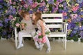 Little twin girls in the garden on a bench against the background of a floral wall with bouquets of flowers in their hands Royalty Free Stock Photo