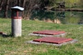 Two locked heavy metal manhole covers mounted on concrete foundation surrounded with concrete ventilation pipe and green grass