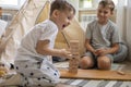 Two male children playing Jenga hardwood bricks tower construction childish room with linen wigwam