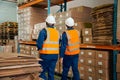 Rear view of two male workers in safety workwear walking near the shelves with stack of goods in warehouse