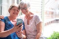 Two mature sisters or friends sitting at home close to the window sharing news on mobile phone. Technology, social media, Royalty Free Stock Photo