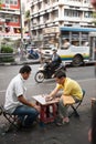 Two men enjoy playing checkers with bottle caps sitting on a noisy street in Bangkok during the day.