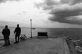 Two men are fishing on concrete pier with fishing rods. There is a bench at the pier. Photo black and white shot and cloudy sky