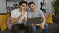 Two men interacting with a laptop on a couch in a home environment Royalty Free Stock Photo