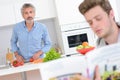 Two men in kitchen one preparing vegetables other reading from recipe book Royalty Free Stock Photo