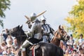 Two men knights riding horses on the battlefield and having a fight on wooden swords - people watching behind the fence