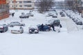 Two men pushing stuck Hyundai Solaris car through a snowy yard between rows of parked cars in deep snow in slope.