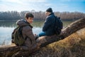 Two men sitting on a log by the river, father and son, early spring landscape, hiking and outdoor activities concept