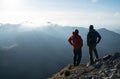 Two men standing standing with trekking poles on cliff edge and looking at sunset rays over the clouds. Successful summit concept Royalty Free Stock Photo