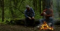 Two men are talking sitting on log in forest near flaming campfire in dusk time Royalty Free Stock Photo