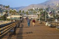 Two men walking along a brown wooden pier at the beach with mountain ranges and blue sky with American flags along the pier