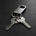 Two metal keys on a ring lie on a dark surface. Above them is a rectangular metallic