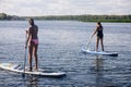 Two middle aged women puddle boarding on beautiful blue lake with oars in hands with trees and reeds in background both