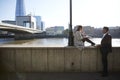 Two millennial colleagues take a break sitting on the embankment eating near London Bridge by the River Thames, backlit Royalty Free Stock Photo