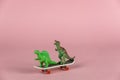 Two miniature dinosaurs riding a mini skateboard. Small green children`s toys against a pink background. Close-up.  Selective Royalty Free Stock Photo