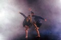 Two modern dancers stretching their shoe-less feet high in the air surrounded by smoke on stage
