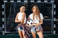 Two modern young teenage hipster girls posing with happy faces in front of goal post at the soccer stadium. Royalty Free Stock Photo