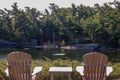 Two Muskoka chairs sitting on a wood dock facing a calm lake. Across the water is a white cottage nestled among green trees