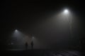 Two mysterious hooded men walk away down a dark foggy road, dimly lit