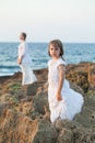 Two nice sisters in white clothes, kid and teenage girl standing on rocks near sea on the background of blue sky Royalty Free Stock Photo