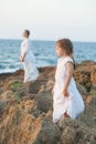 Two nice sisters in white clothes, kid and teenage girl standing on rocks near sea Royalty Free Stock Photo