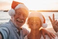 Two old happy seniors wearing christmas hats at the beach taking a selfie of them smiling and having fun with the sunset at the Royalty Free Stock Photo