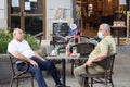 Two Old senior men, friends, one wearing a respiratory face mask, the other not, sittting at the terrace of a bar drinking