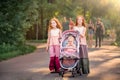 Two older redhead sisters are walking in park on a sunny summer evening. with a stroller with her little sister. Girls in long